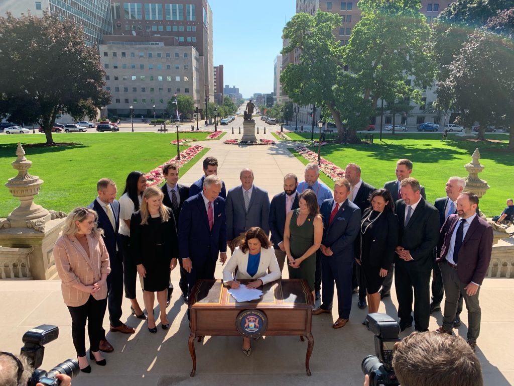 A large group of people stand around Michigan Governor Gretchen Whitmer on the east front steps of the Michigan capitol. Gov. Whitmer is seated at a desk, signing a document.